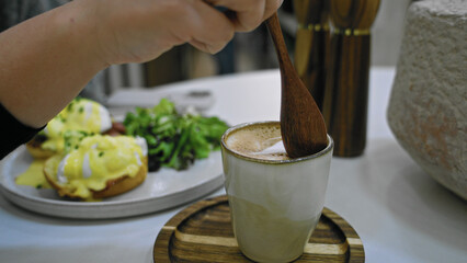 Hand stirring coffee with wooden spoon next to eggs benedict and salad on table in cafeteria