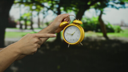 Man pointing at yellow alarm clock held in hand in a bright outdoor park setting during daytime showcasing arms and greenery.