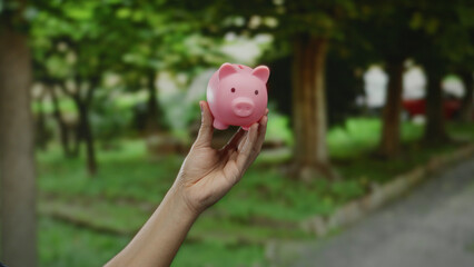 Hand holding pink piggy bank outdoors in a green park setting suggesting savings and financial...