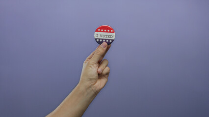 Hand holding a patriotic button reading 'i voted' against a solid purple background symbolizing...