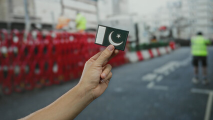 Man holding a small embroidered pakistan flag in city street environment outdoors with traffic barriers in background.