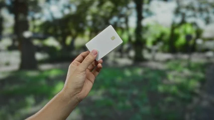 Fotobehang Zomer Man holding white credit card in outdoor park setting with lush green background, showcasing finance and technology concept in a natural environment.  © Krakenimages.com