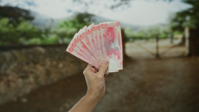 Man holding philippine pesos in an outdoor park setting, showcasing money against a natural backdrop, emphasizing finance and nature contrast.