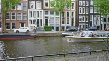 Fototapeta premium Defocused urban view of amsterdam canal with traditional buildings, boats, trees, and cars illustrating local architecture and motion blur in the netherlands.