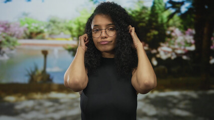 Woman with glasses and curly hair covers her ears in a sunny green park beside a pond and...