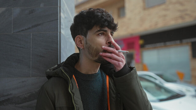 Young man smoking standing in urban street setting, showcasing thoughtful expression, wearing casual winter jacket and sweater with cars and buildings in the background. - Powered by Adobe