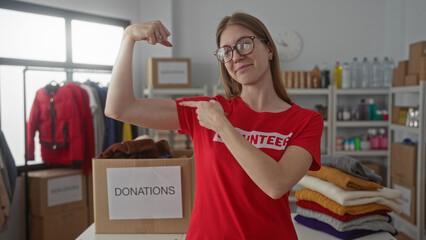 Woman flexes bicep beside cardboard donation box and folded sweaters on table in volunteer center...