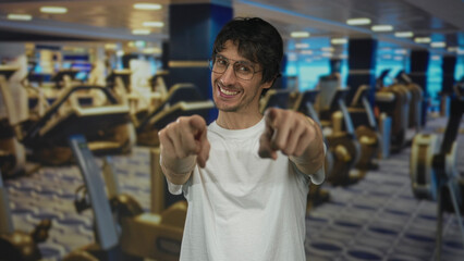 Young man wearing glasses and white shirt points finger at camera in building gym filled with...