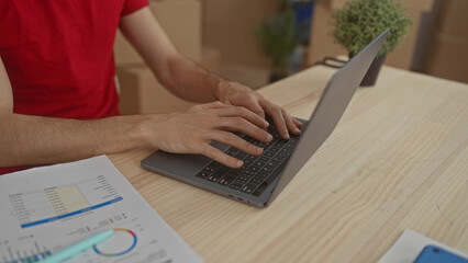 Young man in red shirt typing on laptop at wooden desk in building surrounded by cardboard boxes...