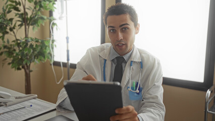Fototapeta premium Young hispanic man doctor with stethoscope in uniform using tablet in a clinic room showcasing a professional indoor healthcare setting.