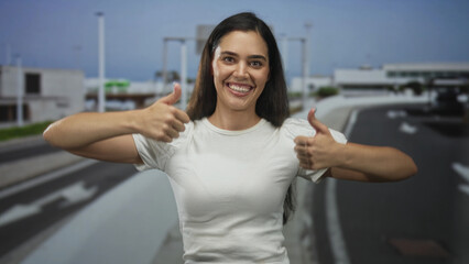 Smiling woman extends hand with thumbs up gesture on highway outdoors under clear sky in daylight;...