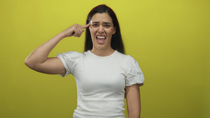 Young hispanic woman in white t shirt points finger to temple on yellow studio set showing tense...