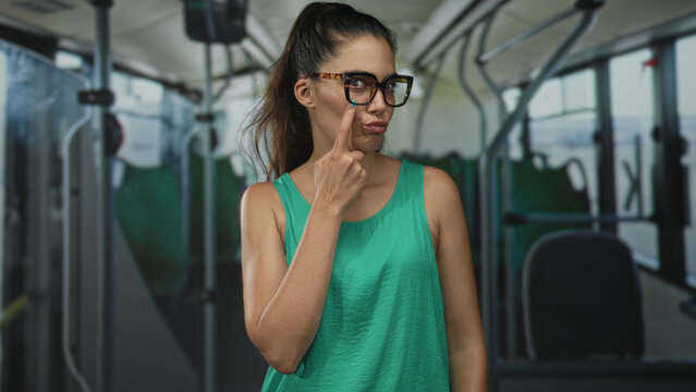 Woman points finger to eye on a bus interior street scene, young hispanic wearing eyeglasses and green tank top; playful attention.