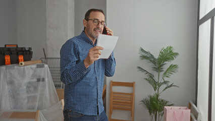 Man calls while reading note in modern living room with unpacked boxes and indoor plant suggesting new home atmosphere.