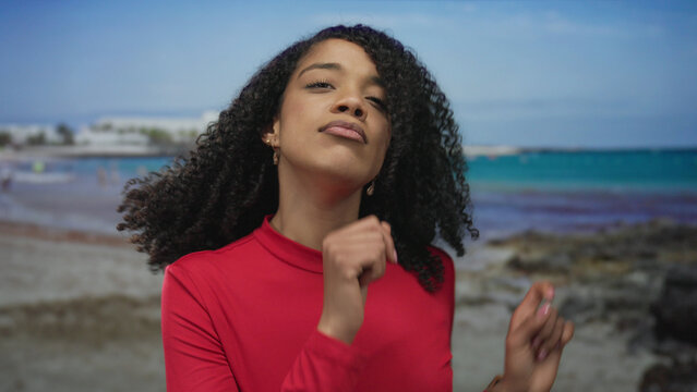 African american woman wearing red top dancing with raised hands at sandy seaside beach in bright daylight; joy fun celebration energy. - Powered by Adobe