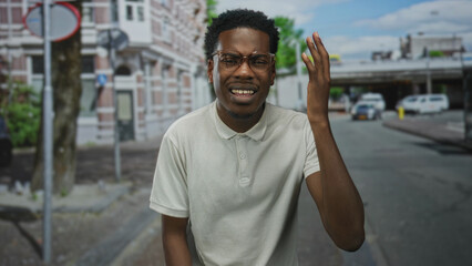 Young african american man wearing glasses raises hand to head on busy city street amid cars;...