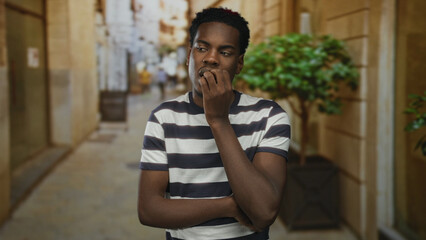 Man in striped shirt bites fingernails on narrow city street lined with stone walls under warm...