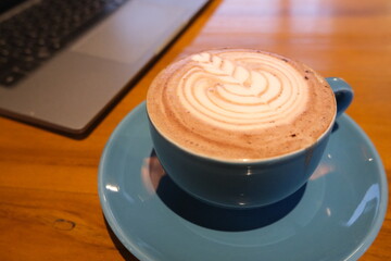 Close-up of a creamy cappuccino topped with beautiful latte art, served in a blue ceramic cup and saucer on a warm wooden surface.  
