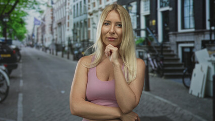 Woman with hand on chin standing on a city street in daylight, gazing thoughtfully at the camera;...
