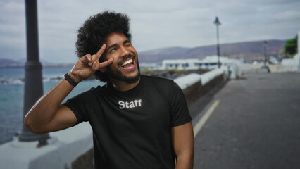 African american man in staff uniform makes victory gesture outdoors on seaside promenade, smiling brightly under cloudy skies with serene sea view.