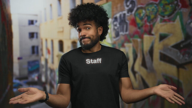 Man in staff uniform with afro shrugging on graffiti street background outdoors in city environment expressing confusion with urban art visible. - Powered by Adobe