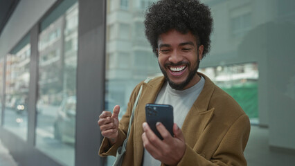 Man celebrating success on smartphone in city street with joyful expression outdoors in urban setting wearing casual jacket and smiling broadly.