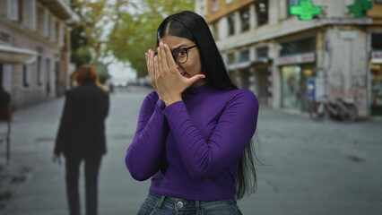 Young latin woman standing covers face with hands on city street under pharmacy cross sign;...