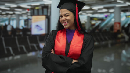 Woman wearing graduation cap and gown stands with arms crossed in airport terminal; success...