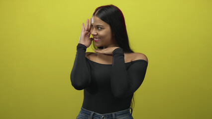 Woman framing her face with one hand on temple and one hand under chin in a yellow studio;...