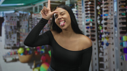 Young latin woman sticks out tongue and makes l sign at colorful souvenir market stall; playful...