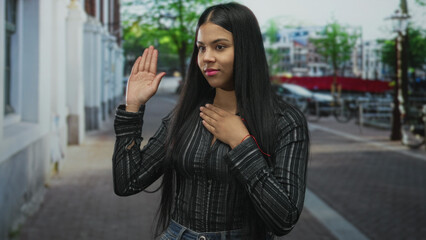 Woman raises right hand and presses left hand to chest on street with focused expression; honesty...