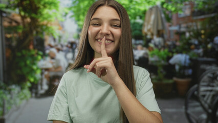 Teenage girl finger to lips for silence at outdoor restaurant terrace under sunlit leafy canopy...