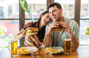 Happy couple eating burger sitting at pub restaurant fast food table - Young people having lunch break at cafe bar venue - Life style concept with guy and girl hanging out on weekend day