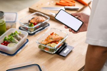 Man holding mobile phone with white screen mockup over healthy meal prep for weight loss at home.