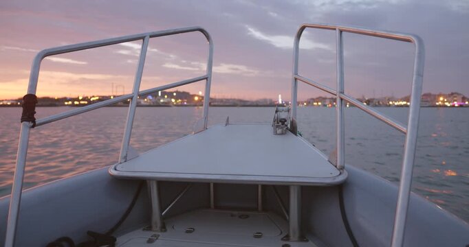 Cinematic POV from the front of a boat at dawn in Agde, France, with city lights reflecting over calm waters under a colorful morning sky.