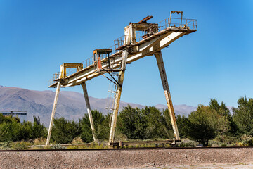 An old rusty abandoned gantry crane standing on rails, bushes and mountains in the background.