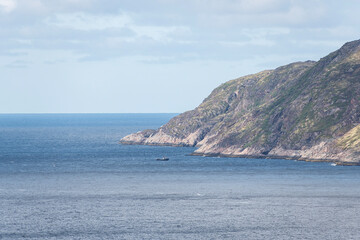 A small boat glides through calm waters beside a rocky shoreline, with lush green hills rising in the background under a bright blue sky.