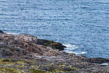 A rugged coastline reveals large rocks and scattered vegetation near the water, where gentle waves lap against the shore under bright midday light.