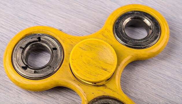 A yellow fidget spinner with three metallic bearings rests on a light wooden surface—evoking tactile focus, playful motion, and the everyday simplicity of stress-relief tools.