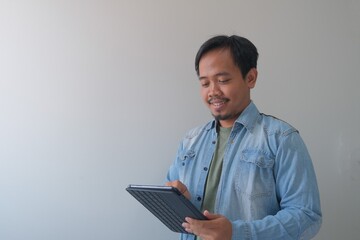 Happy man in denim jacket looking at a tablet device, standing against a plain wall in a bright...