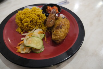 Hearty meal of yellow spiced rice served with grilled chicken, pickled vegetables, and sambal on a red and black plate.