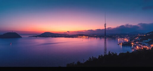 Calm waterfront cityscape at dusk with illuminated buildings, calm sea, surrounding hills, and a tall communication tower under a vibrant twilight sky