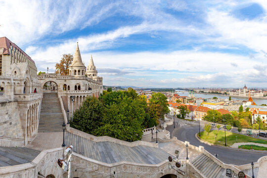 Wide panorama of Fisherman's Bastion with conical towers, arcades, and grand staircase leading to Danube, featuring Parliament building view under autumn sky. Budapest, Hungary.

