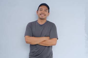 A cheerful man wearing a gray shirt and cap stands against a plain background with his arms...