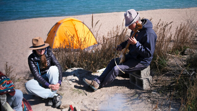A man plays a national Kyrgyz musical instrument - komuz or Kyl Kyyak. A loving father spends time with his adult daughters on the beach by the sea