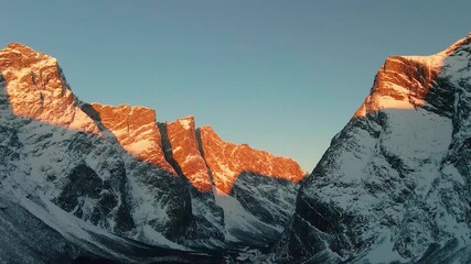 This drone footage shows beauty of the Troll Wall Trollveggen in Norway, captured during the golden hour as the setting sun casts a warm alpenglow across its snow-capped peaks. - Powered by Adobe