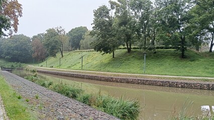 River with embankment and green bank in the park, trees in the background.