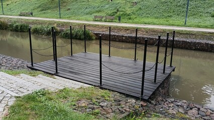 A wooden dock with metal railings and chains on a river or canal. A small pier near the shore,...