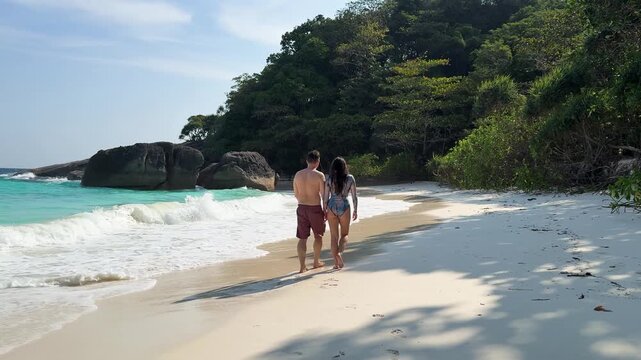Couple walking hand in hand along tropical sandy beach with turquoise water at Similan Islands, Thailand.