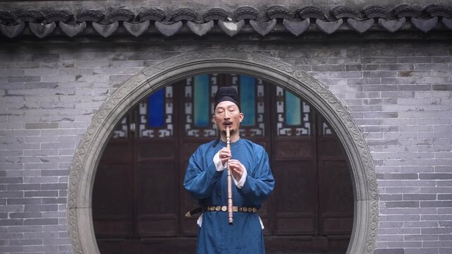 A middle-aged man in traditional blue robe playing bamboo flute in front of an arched gateway in a Chinese courtyard
